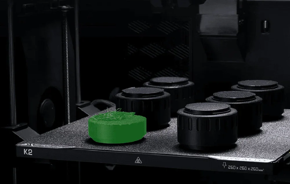 3D printeris ar zaļu modeli uz drukas platformas blakus melniem regulatoriem