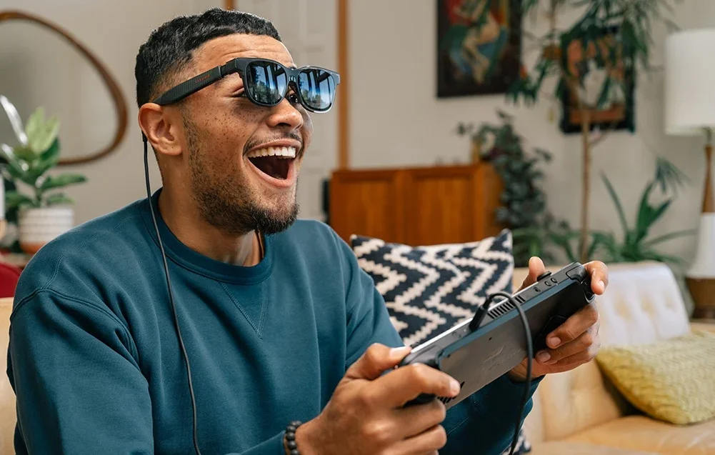 Vīrietis ar XR brillēm smejas, spēlējot videospēles ar kontrolieri rokās viesistabā