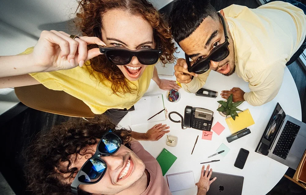 Trīs darbinieki ar XR brillēm smaida pie galda ar klēpjdatoriem un līmlapiņām