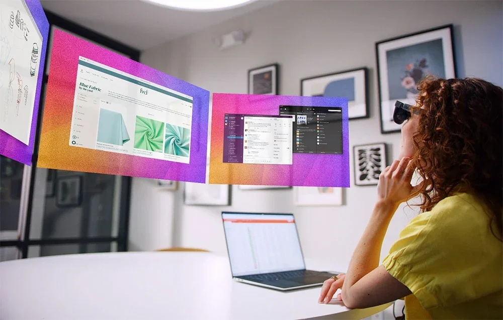 Sieviete ar XR brillēm strādā pie galda ar klēpjdatoru, skatoties virtuālos ekrānus gaisā