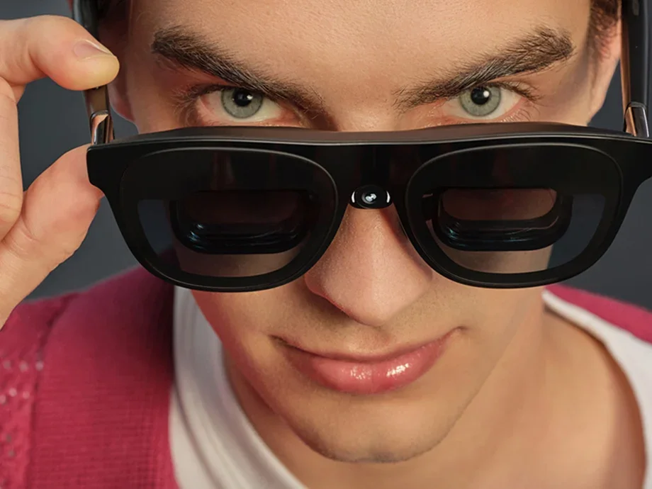 A man approaching the camera, lifting black AR glasses with a camera and looking straight into the lens.