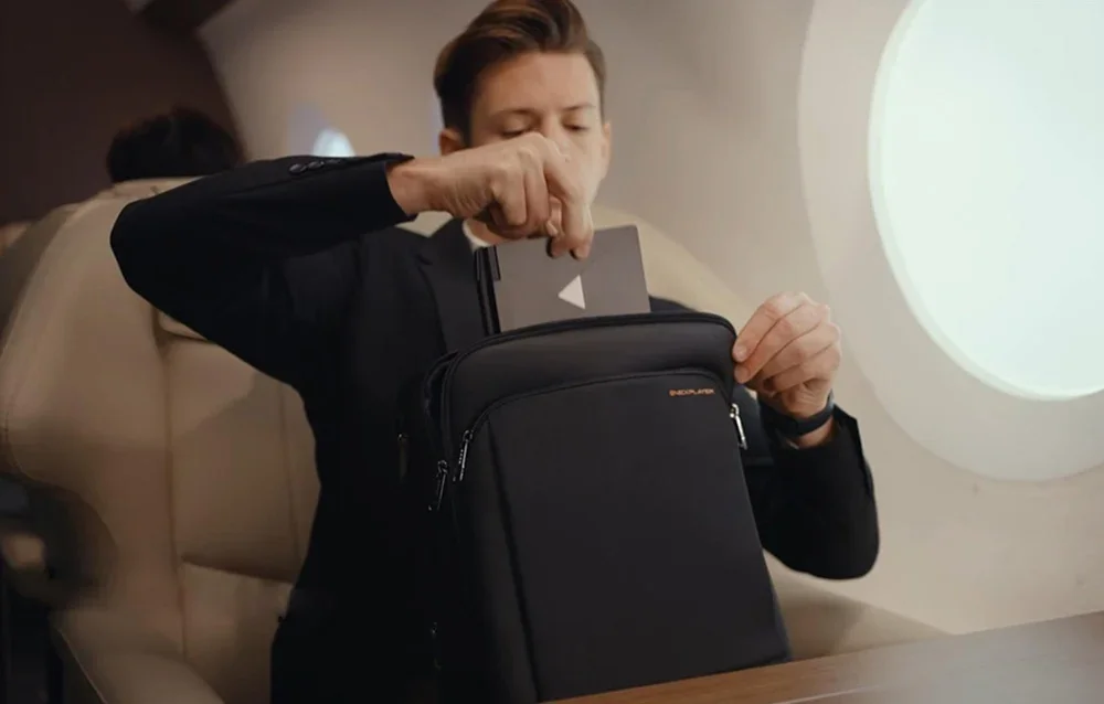 A man in a black sweatshirt puts a laptop into a black travel bag on a plane next to a window