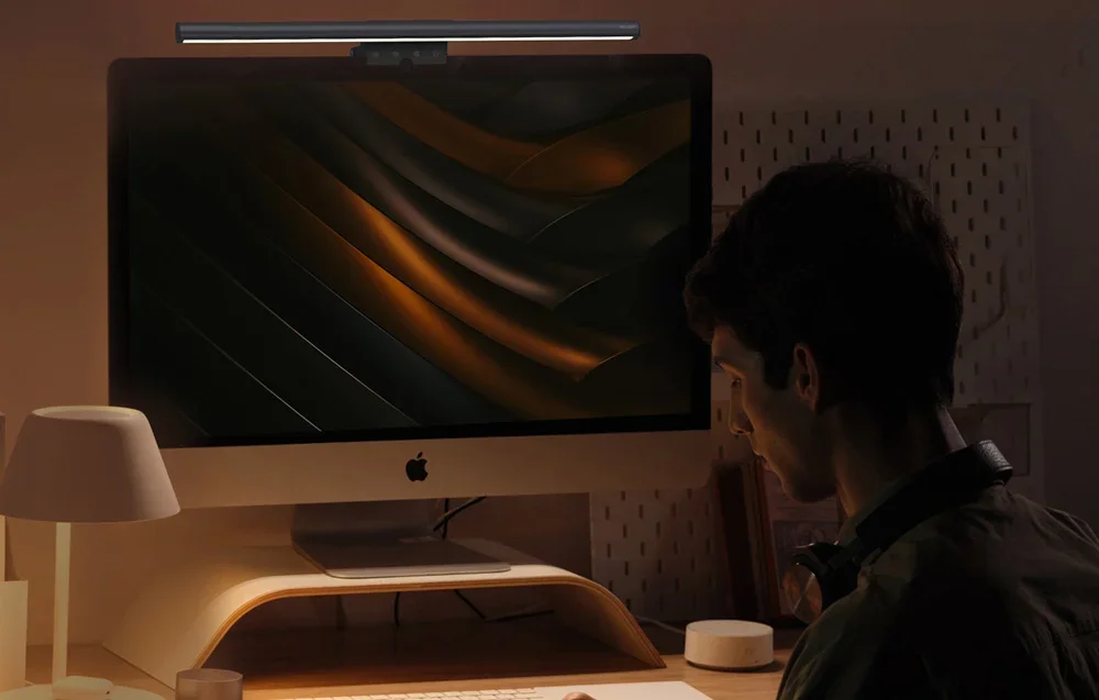 A man sitting in front of an iMac with a lamp mounted on top of the screen, in a dark room