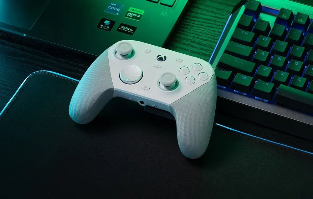 Balts GameSir kontrolieris uz paliktņa blakus klēpjdatoram un tastatūrai.