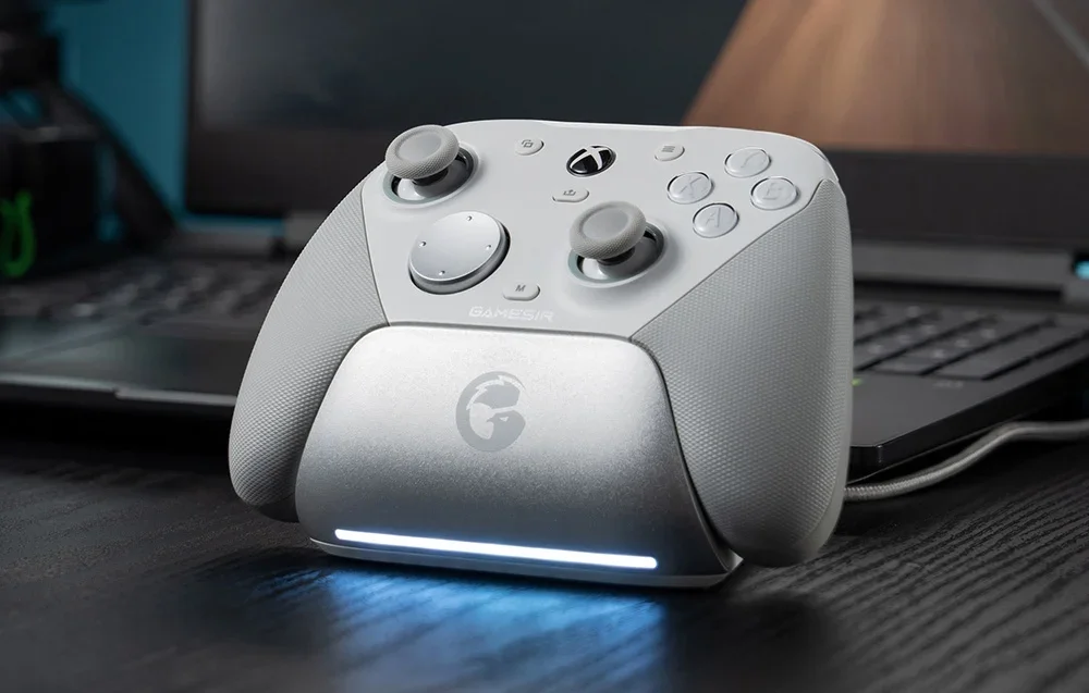Balts GameSir kontrolieris uzlādes stacijā klēpjdatora priekšā.