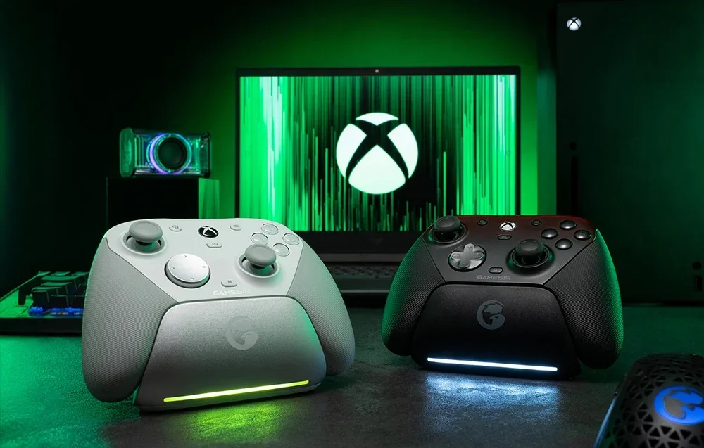 Divi GameSir kontrolieri uz dokstacijas monitora priekšā ar Xbox logotipu, viens balts un otrs melns.